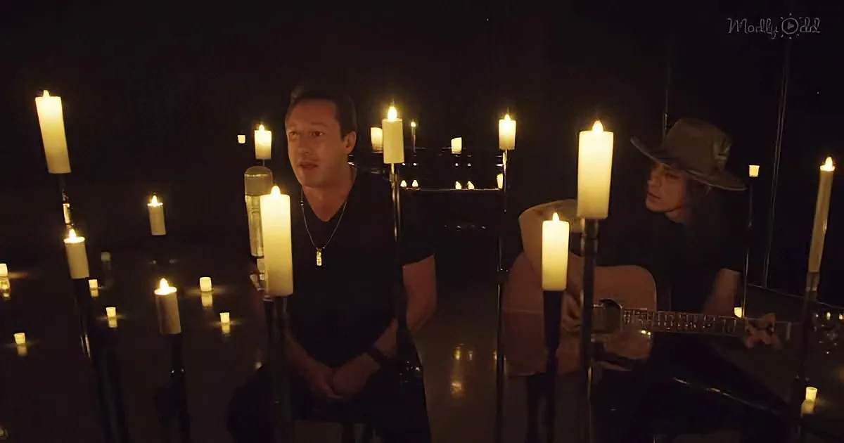 A man in a dark room surrounded by candles, another person playing guitar nearby.