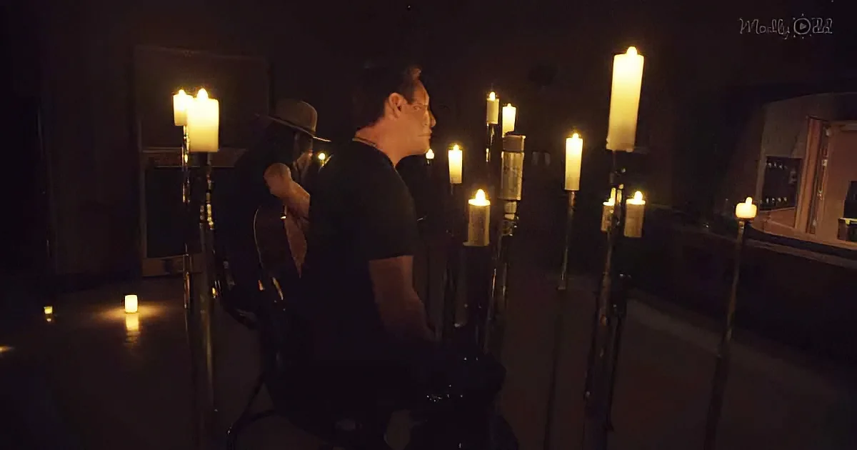 Two people sit surrounded by candles in a dimly lit room, one playing guitar.