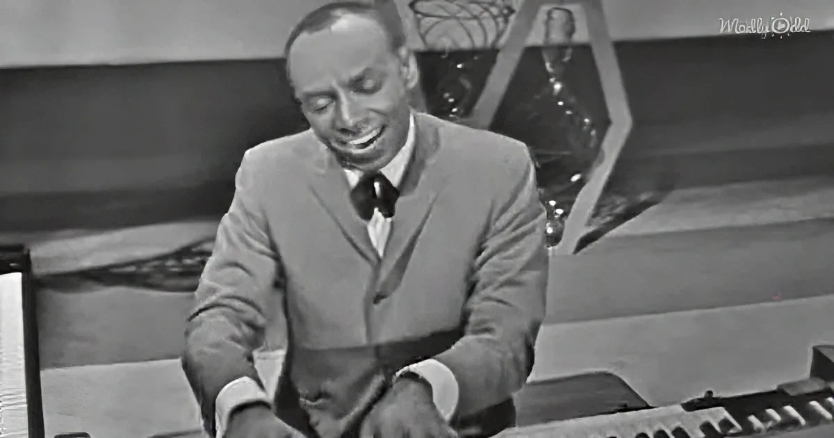 A man in a suit smiles while seated at a desk with a microphone and papers.