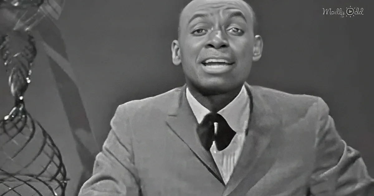 A man in a suit and tie is smiling, with a lamp and a dark background behind him.