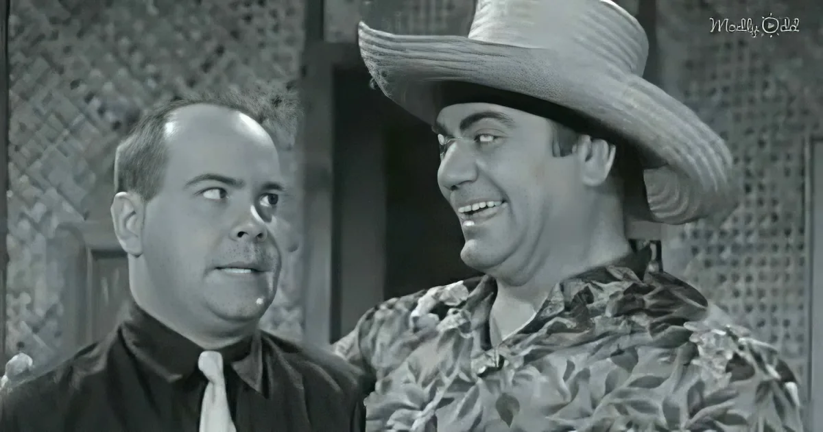 A man in a straw hat and floral shirt smiles while another man in a suit looks up at him.