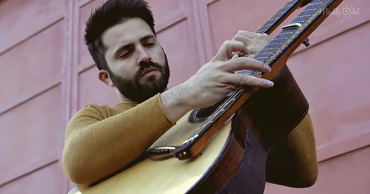 A man with a beard plays a classical guitar against a pink wall.