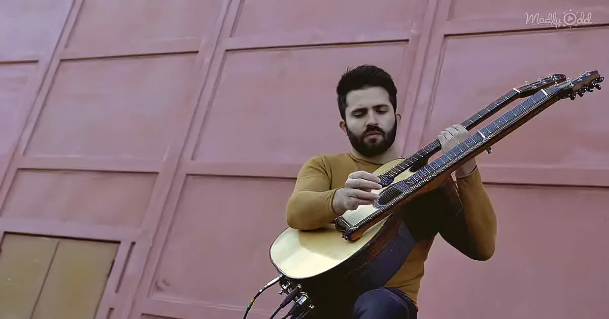 A man with a beard plays a guitar against a pink wall.