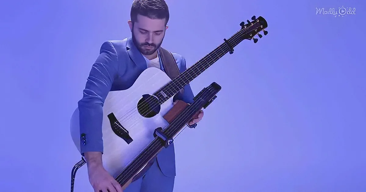 A man in a blue suit plays a white acoustic guitar against a purple background.