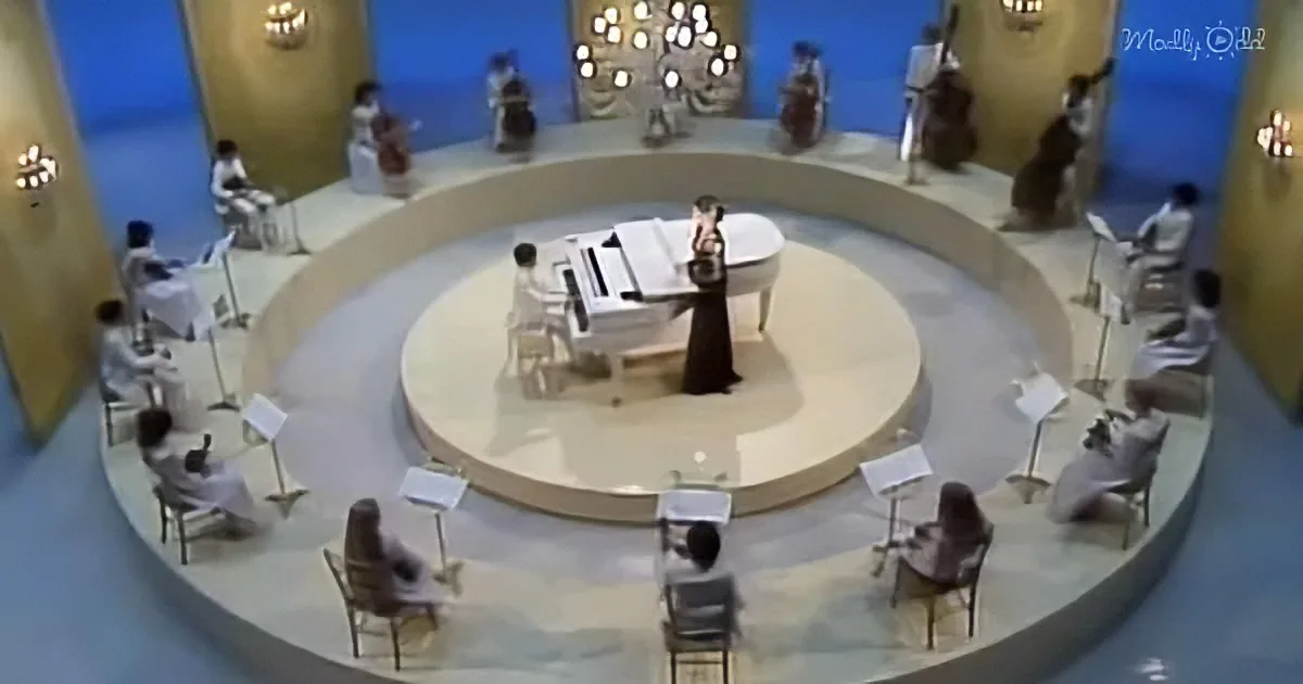 A woman in a black dress stands at a piano on a circular stage surrounded by musicians.