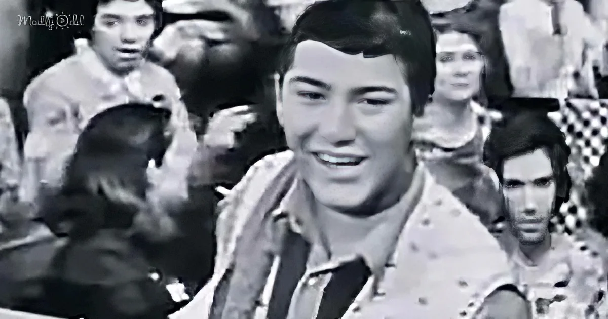 A smiling young man in a patterned shirt, surrounded by a crowd of people, in a black-and-white image.