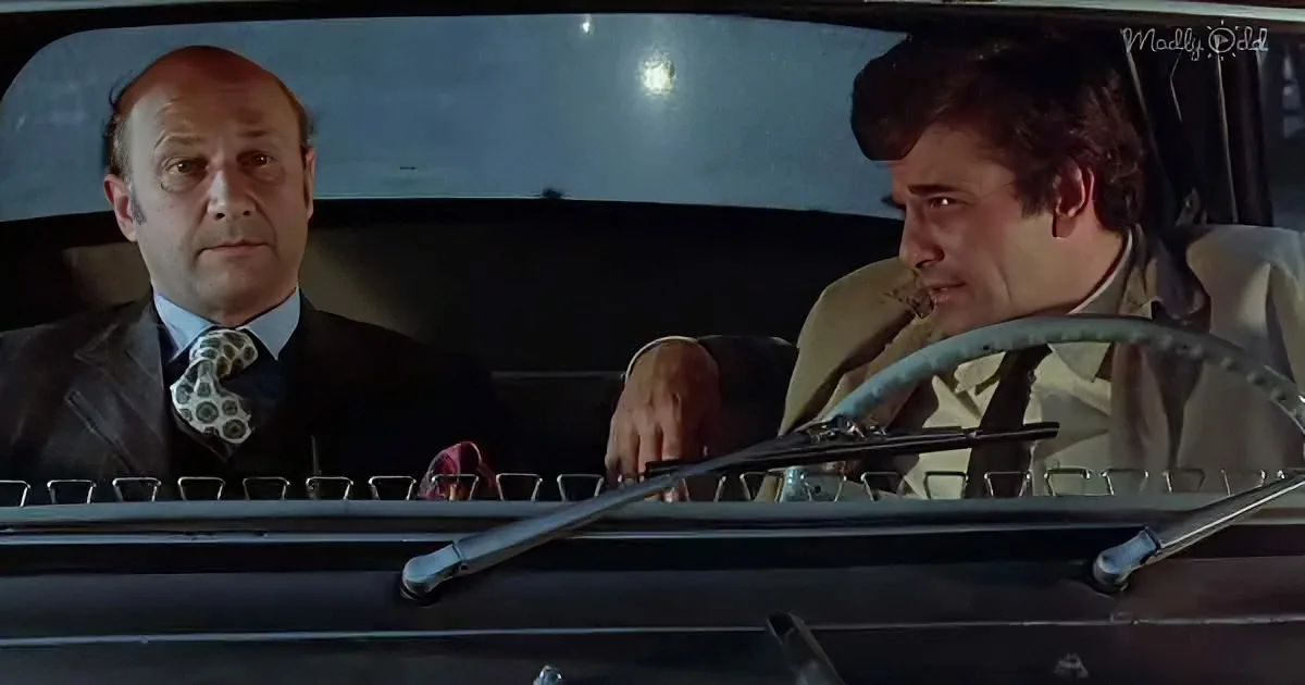 A man in a suit sits in a car, looking ahead, while another man in a tan jacket drives, looking back.