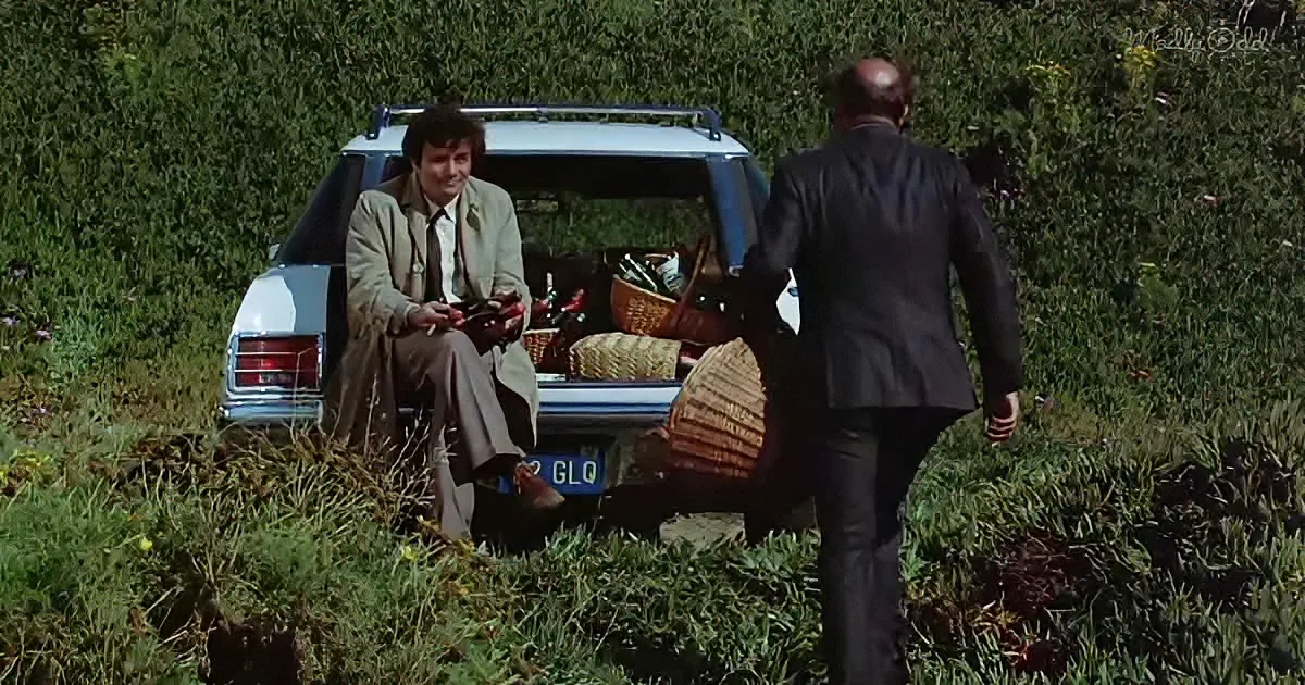 A man in a suit sits on a car's trunk, holding a phone, while another man in a suit walks away.