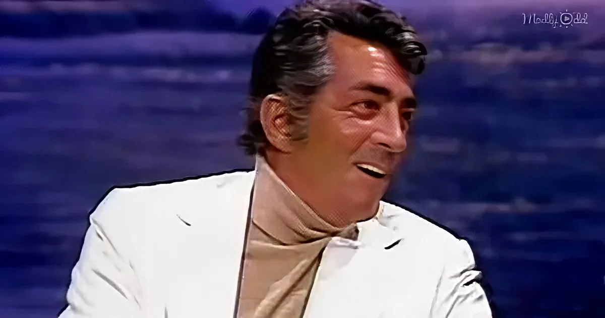 A man in a white suit and beige turtleneck smiles against a blue background.