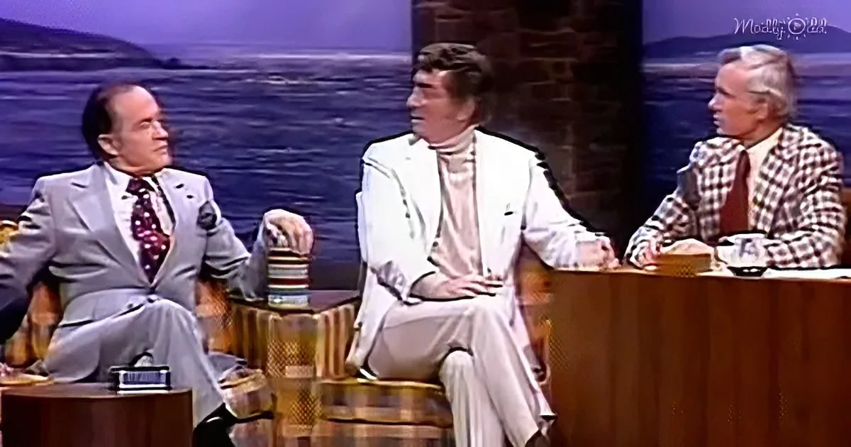 Three men in suits sit at a table, engaged in conversation on a talk show set.