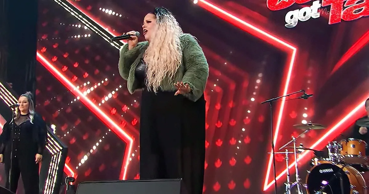 A woman with long, curly hair sings passionately on stage, illuminated by red lights.