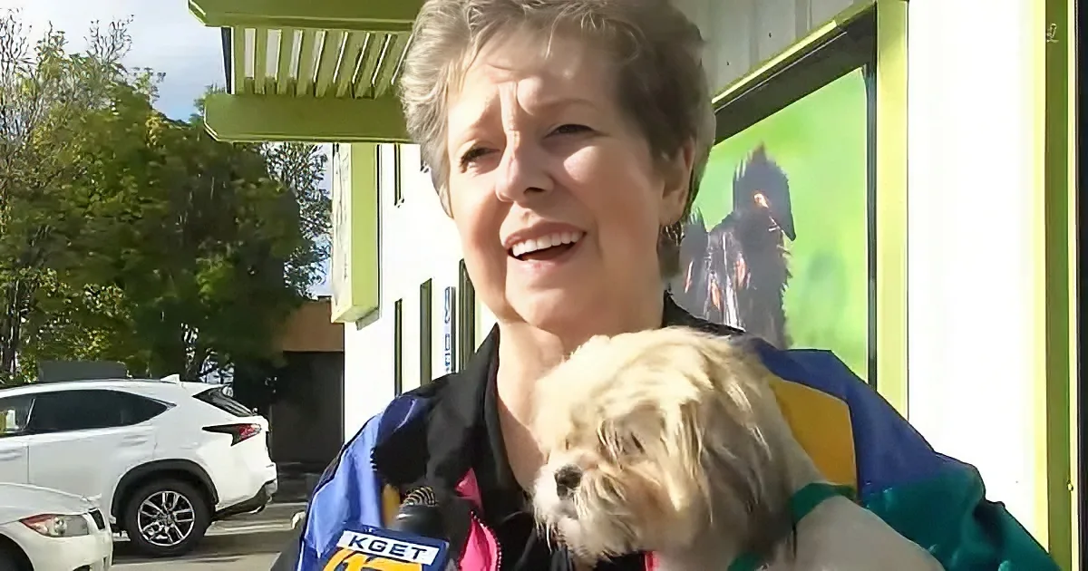 A woman smiles while holding a small dog, wearing a colorful jacket, with a car and building in the background.