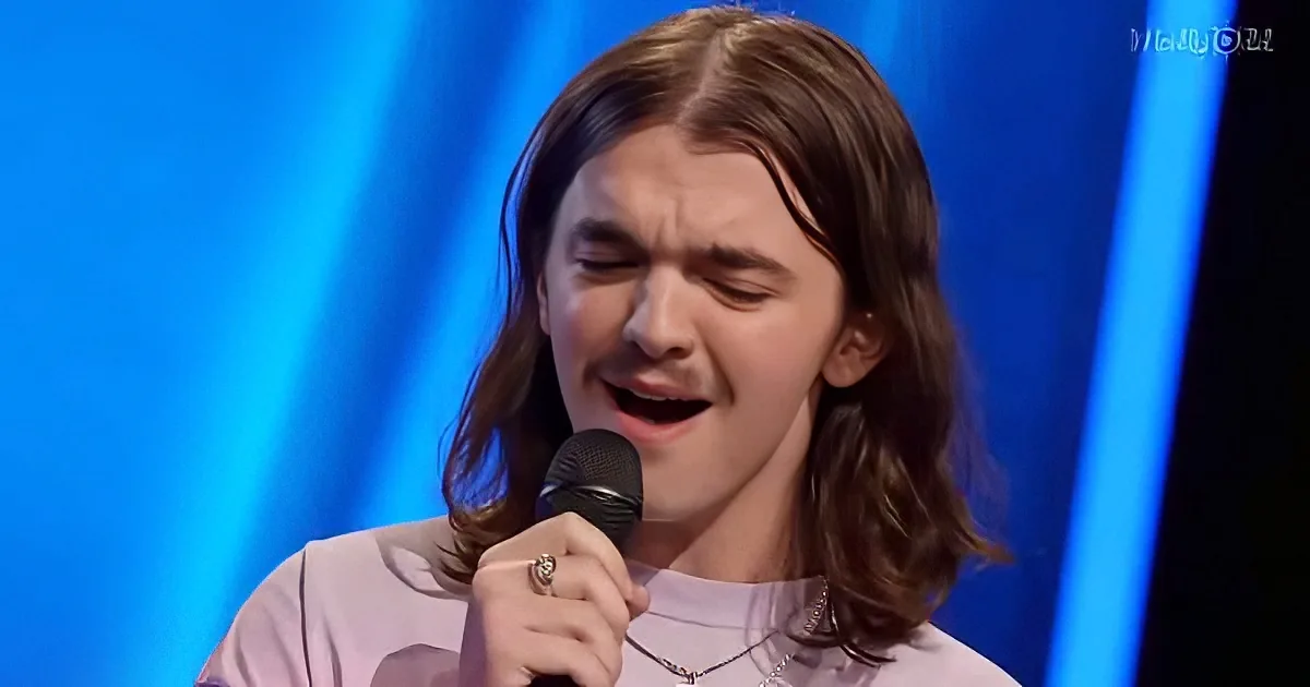 A person with long hair and a nose ring is singing into a microphone, wearing a light shirt and a necklace.