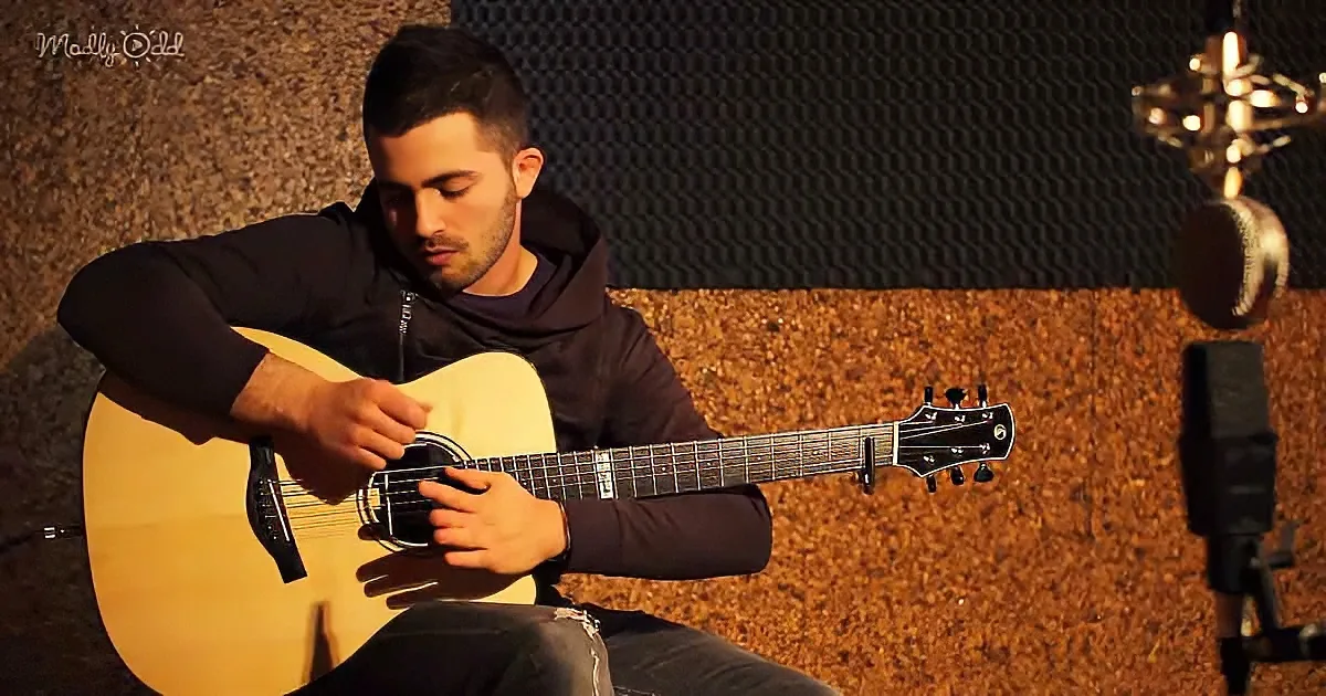 A man in a hoodie plays an acoustic guitar in a dimly lit room with soundproofing panels.