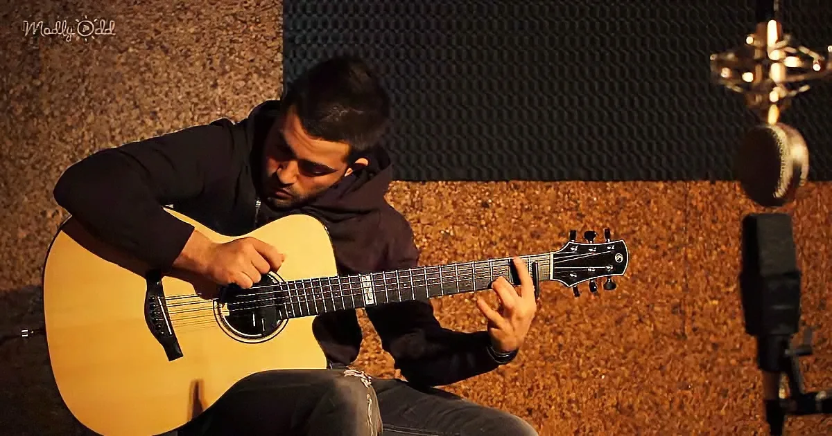 A man in a hoodie plays an acoustic guitar in a recording studio with soundproofing panels.