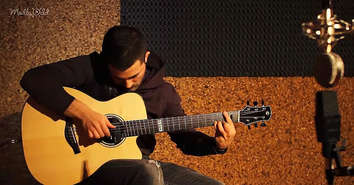 A man sits playing an acoustic guitar in a dimly lit room with soundproofing panels.