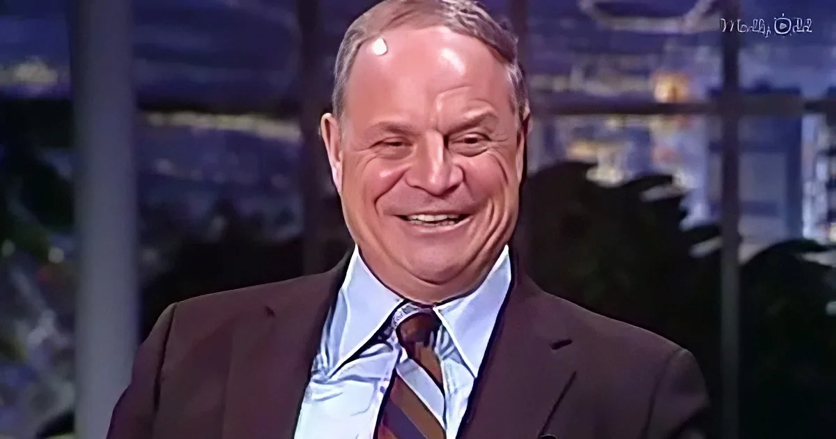 A smiling man in a suit and tie, wearing a blue shirt and brown striped tie, stands in front of a blurred cityscape...