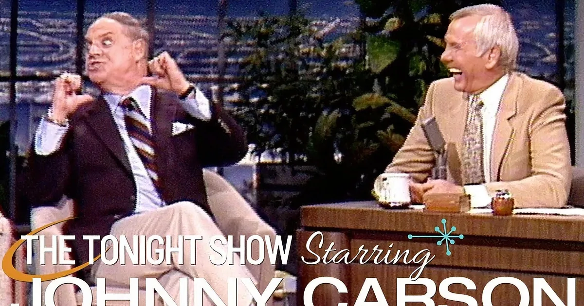 Two men in suits are seated, one in a dark suit with a striped tie, the other in a beige suit with a patterned tie...