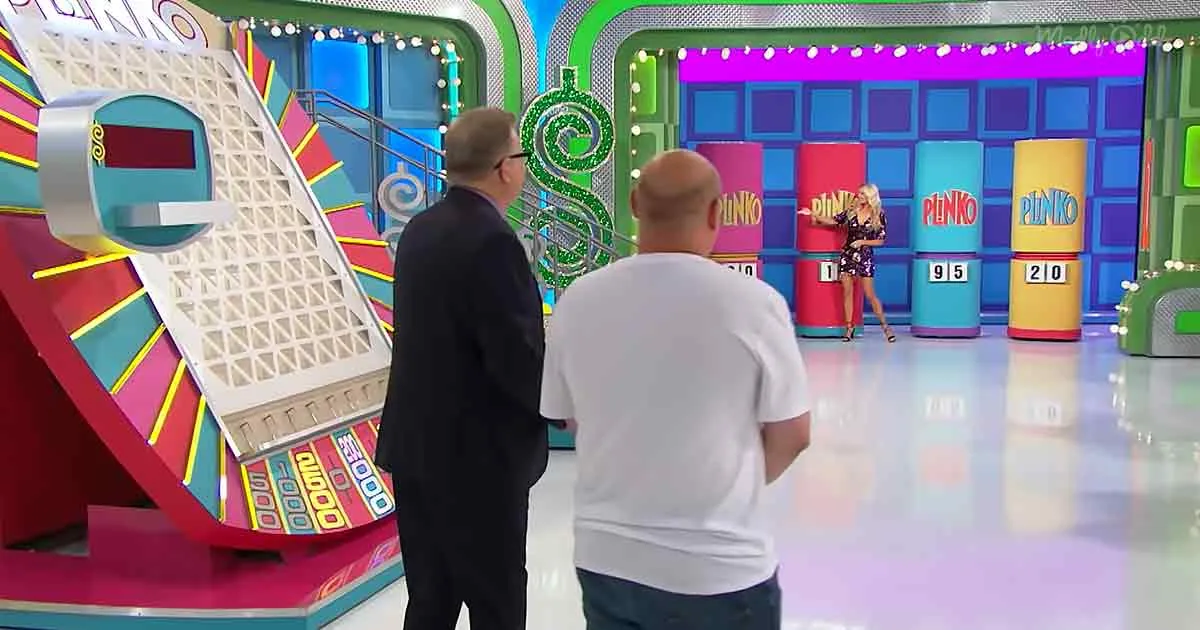 A woman stands on a stage with numbered columns, while two men observe a large, colorful game setup.