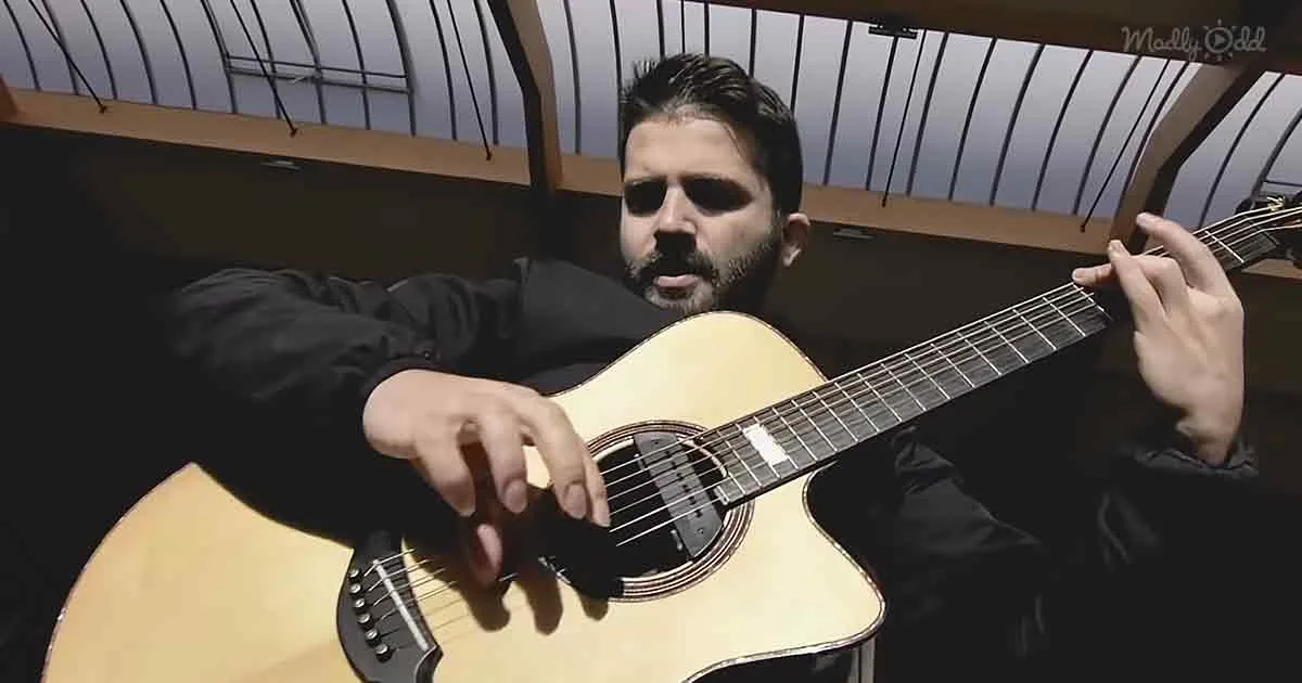 A man with a beard plays an acoustic guitar, wearing a black jacket, in a dimly lit room with a curved ceiling.