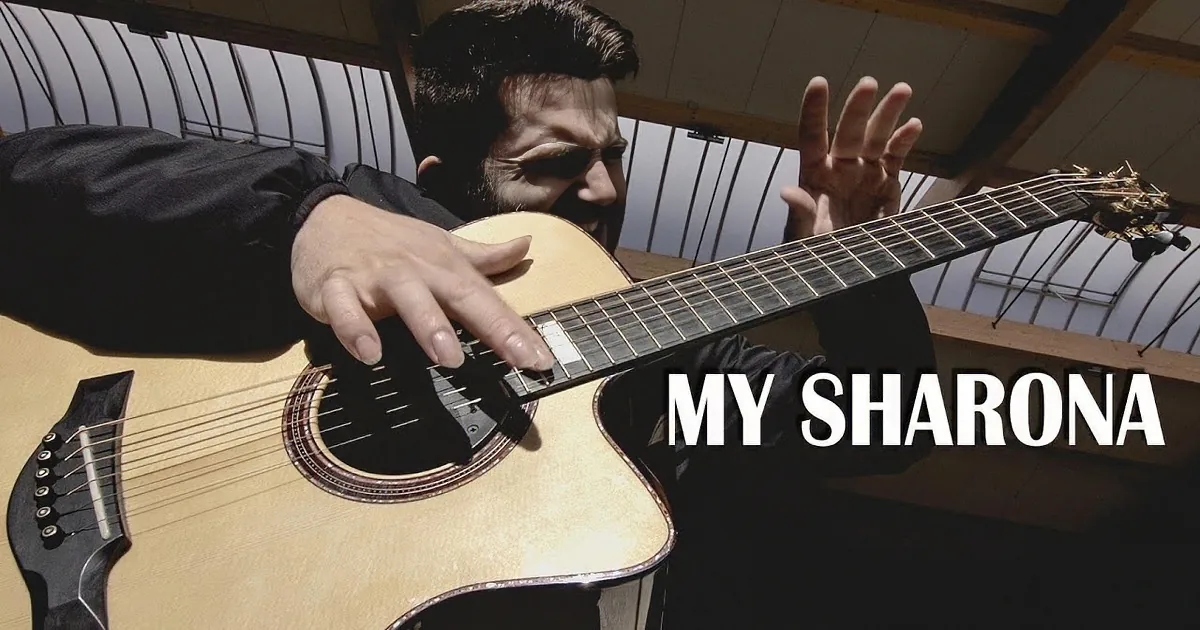 A man in a black shirt plays an acoustic guitar with his left hand on the fretboard and right hand strumming.