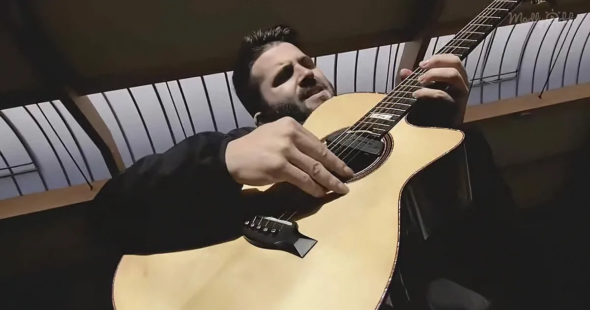 A man in a black shirt plays an acoustic guitar with a windowed background.