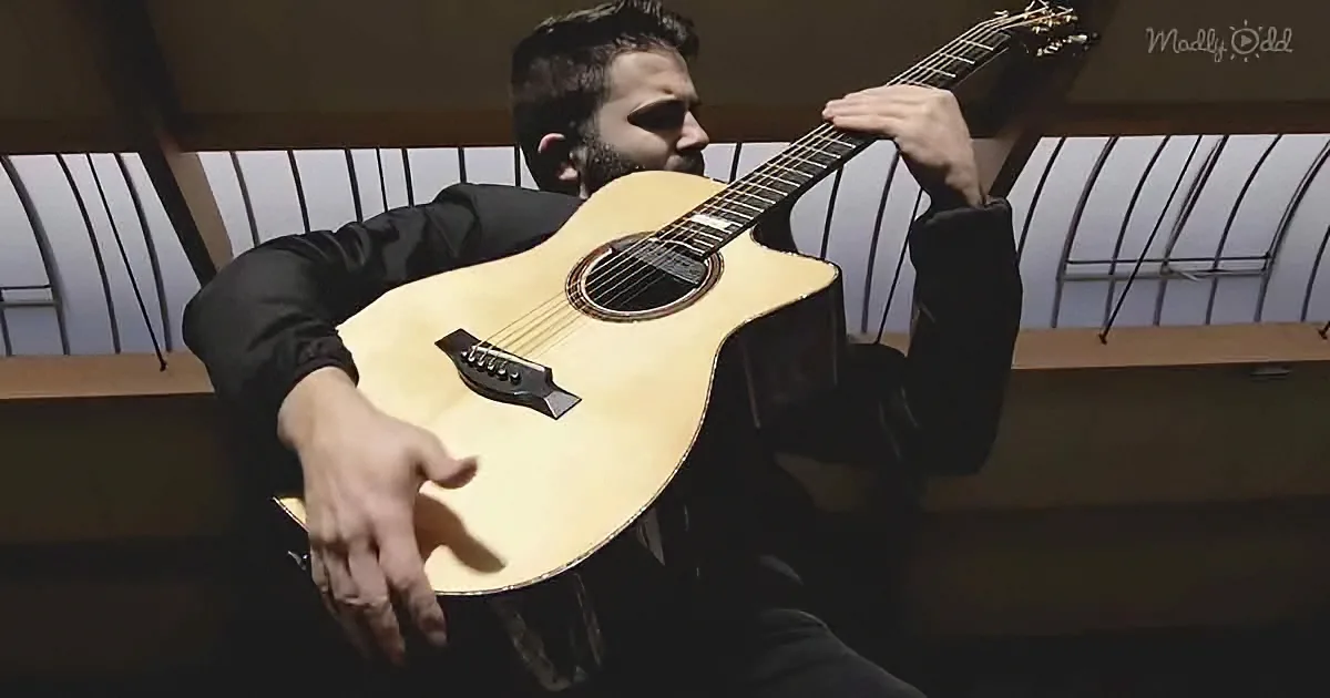 A man in a black suit holds a light brown acoustic guitar, positioned in a dimly lit indoor setting.