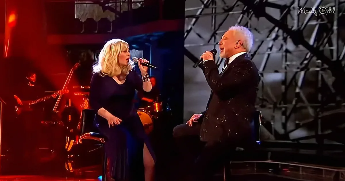 A woman in a blue dress sings into a microphone, accompanied by a man in a suit on stage.