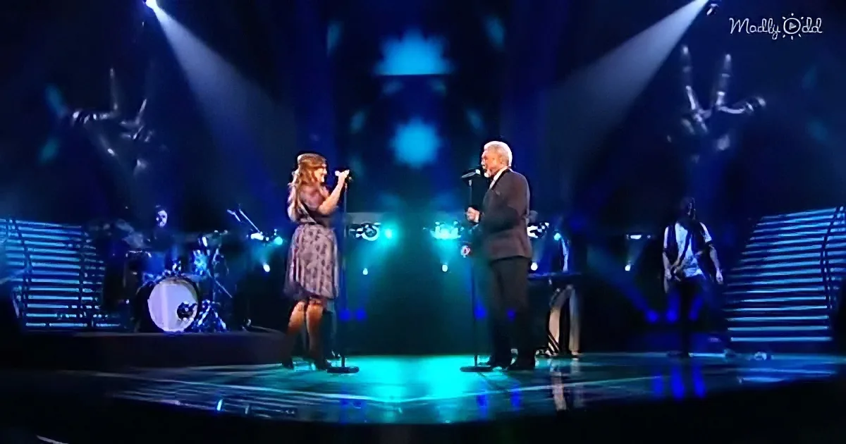 A woman and man sing on stage with a band, blue lights illuminate the scene.