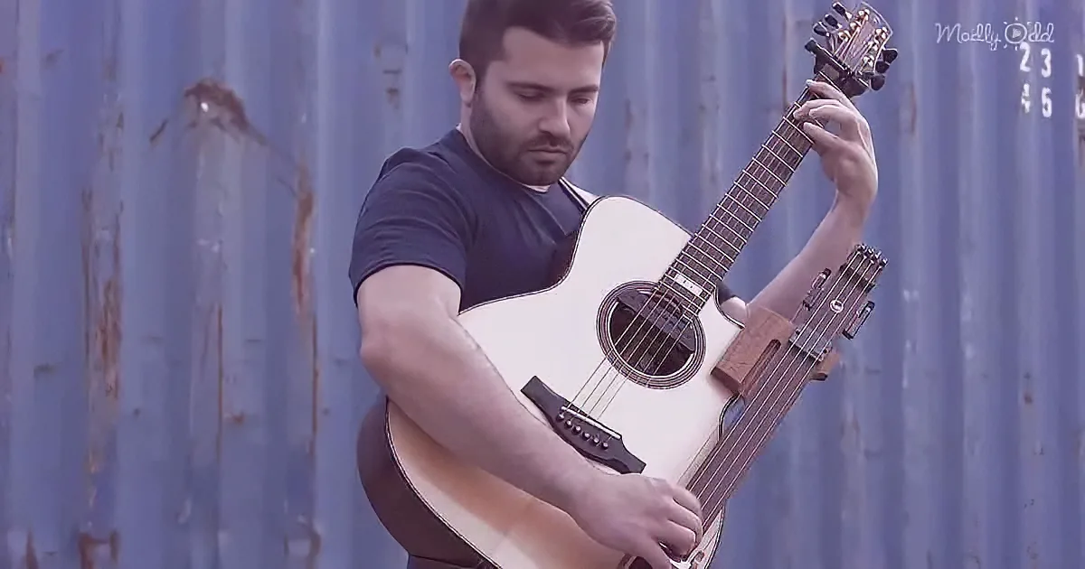 A man with a beard plays an acoustic guitar against a blue corrugated metal backdrop.