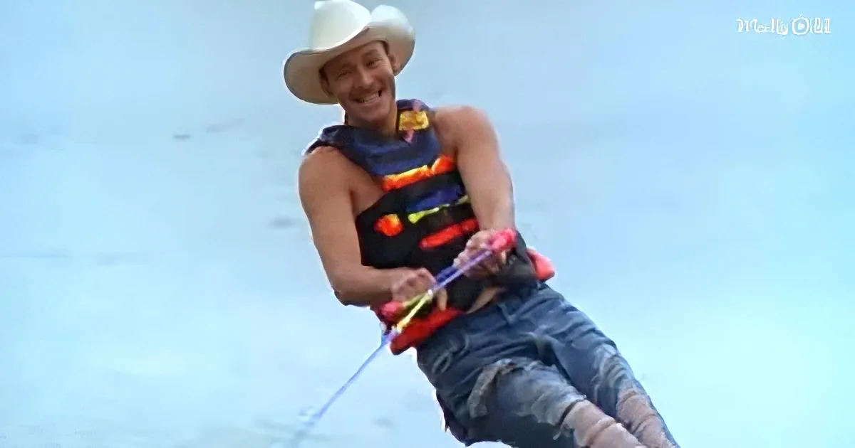 A man in a cowboy hat and life vest is wakeboarding, holding a rope, with a clear sky in the background.