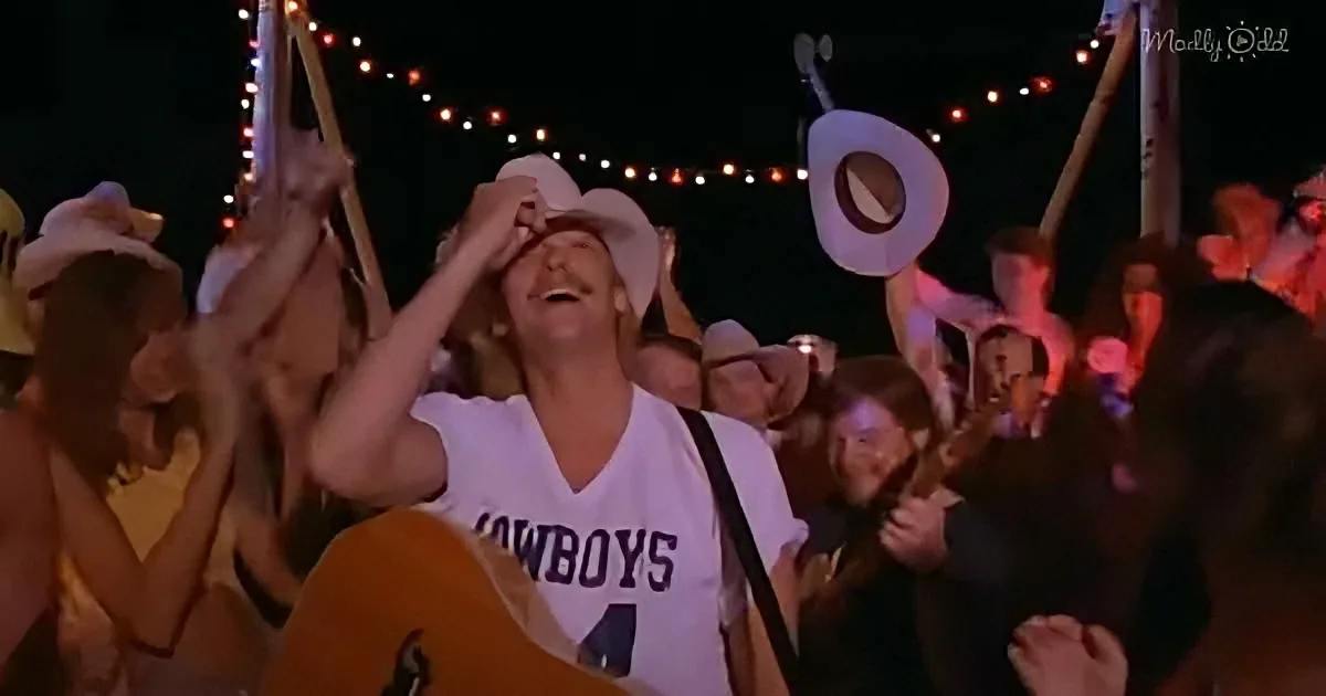 A man in a cowboy hat and "Cowboys" jersey holds a guitar, surrounded by cheering fans at a nighttime event.