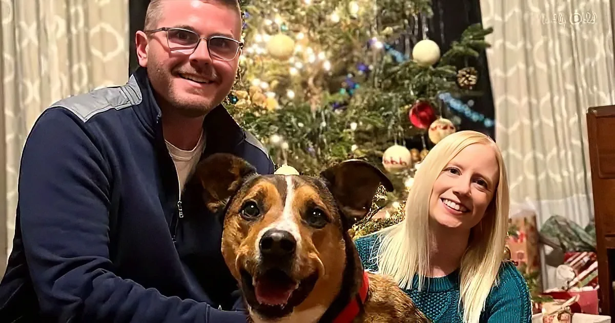 A man and woman smile with a dog in front of a decorated Christmas tree.
