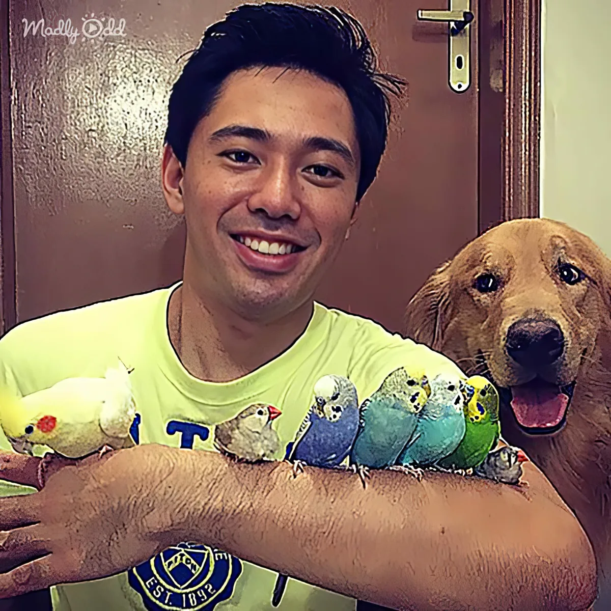 A man smiles with a dog and several colorful birds perched on his arm.
