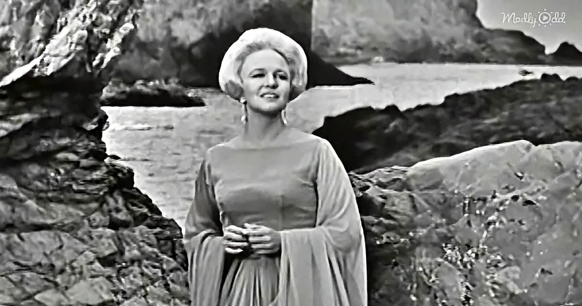 A woman in a flowing dress and large hat stands amidst rocky terrain, smiling.