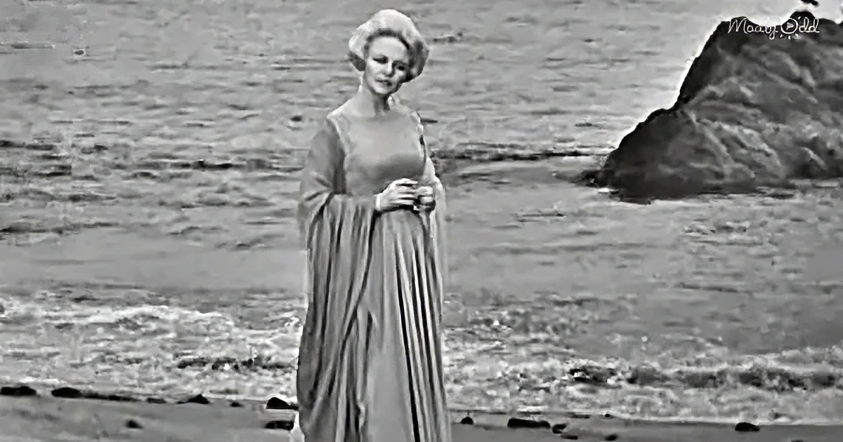 A woman in a flowing dress stands on a beach, holding her skirt, with a rocky outcrop in the background.