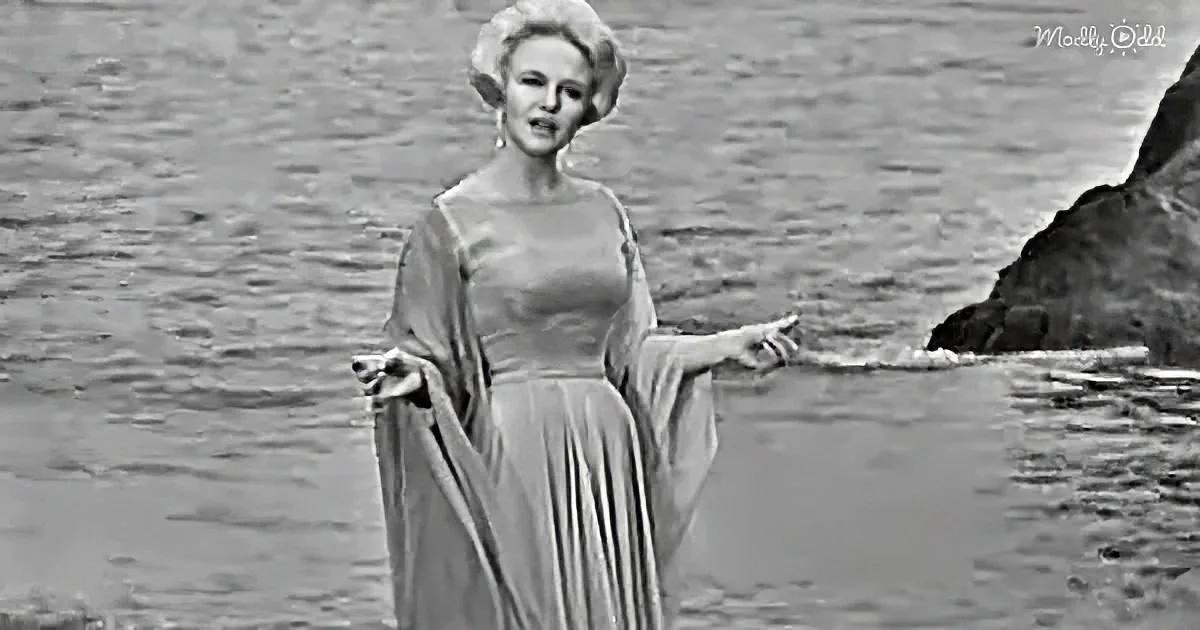 A woman in a flowing dress stands by water, arms outstretched, in a dramatic pose.