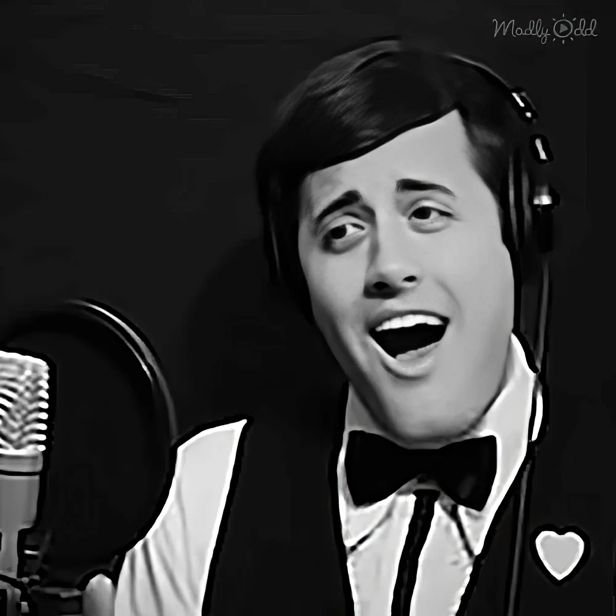 A person in a tuxedo sings into a microphone, wearing headphones, with a dark background.