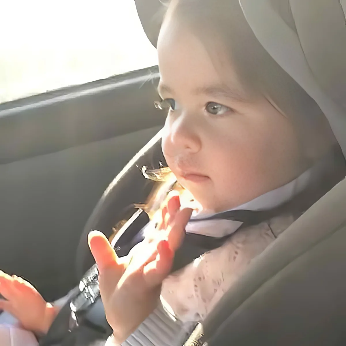 A baby in a car seat, wearing a white outfit, with hands raised, looking to the side.