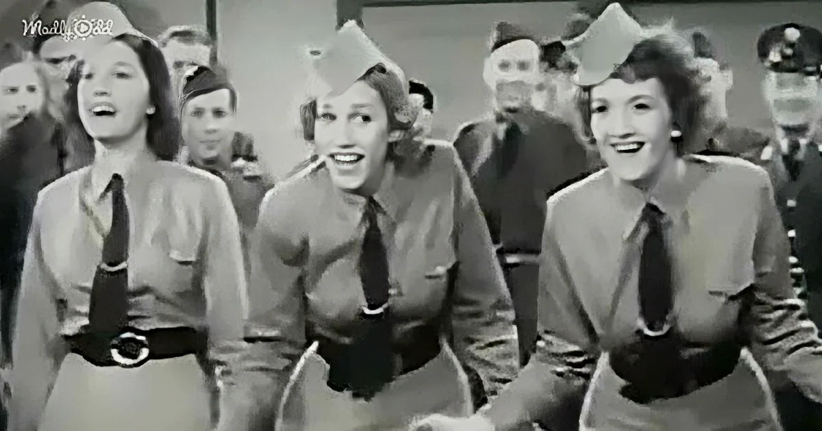 Three women in uniform sing and smile, surrounded by others in military attire.