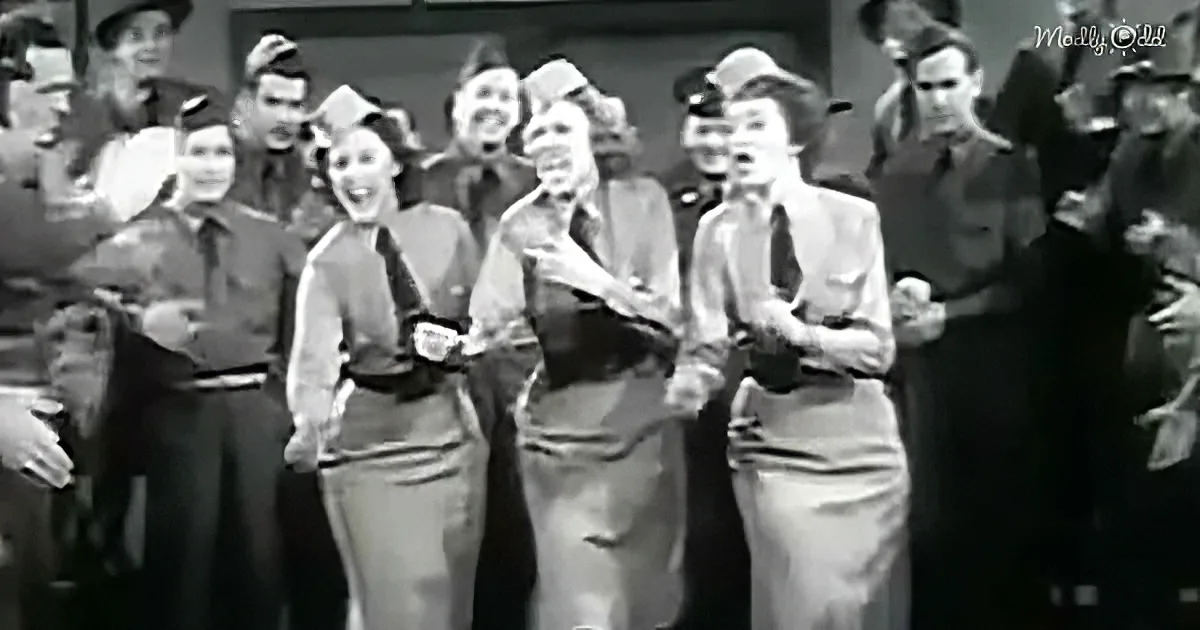 Women in uniform sing, holding sheet music, surrounded by men in military attire.