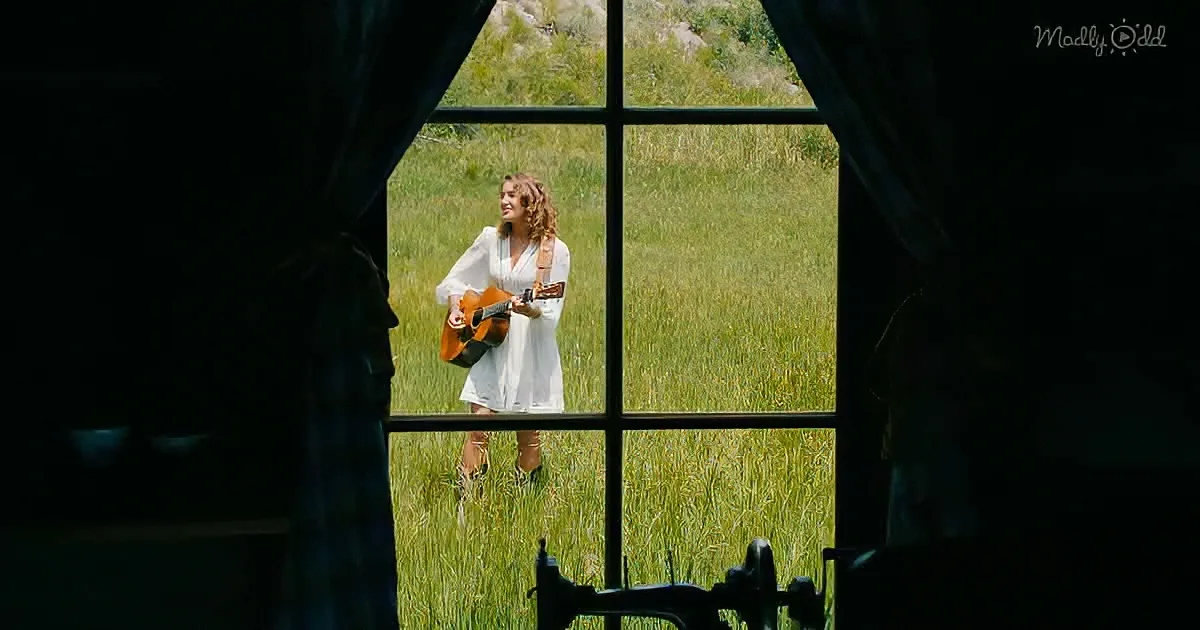 A woman in a white dress stands in a grassy field, holding a guitar, viewed through a window with a sewing machine in the...