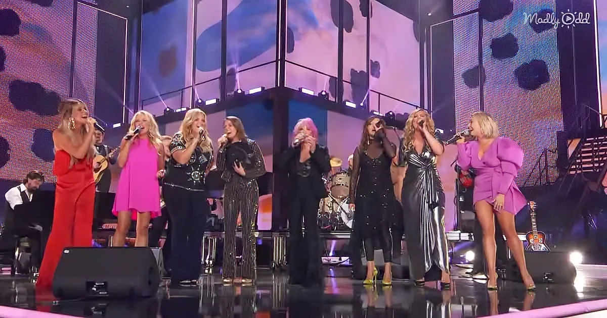 A group of nine women in colorful outfits perform on stage with microphones and a band.