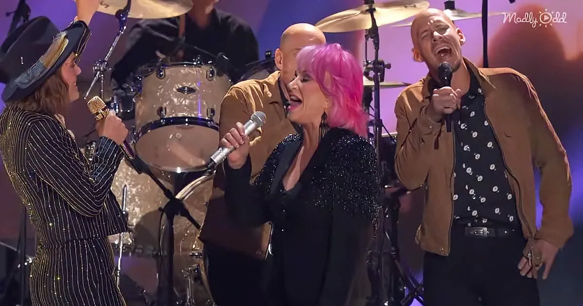 A woman with pink hair sings passionately on stage with a band, surrounded by musicians.