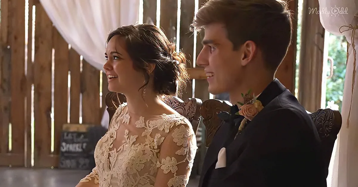 A bride in a lace dress and groom in a black suit smile at each other in a rustic barn setting.
