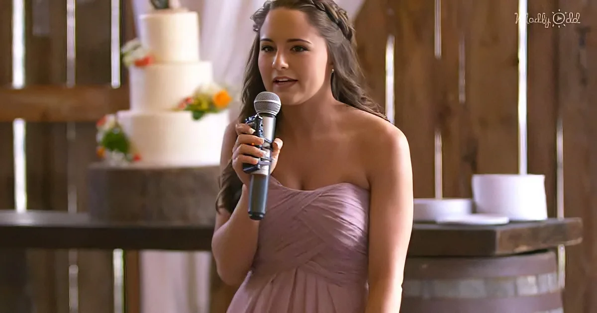 A woman in a lavender dress holds a microphone, standing in front of a tiered wedding cake.