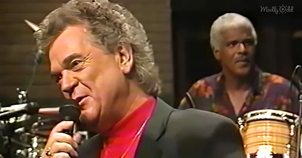 A man in a red shirt sings into a microphone, with another person playing a drum in the background.