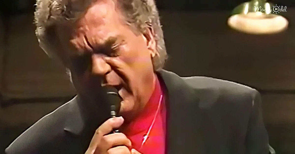 A man in a suit and red shirt sings into a microphone, holding it with his right hand.