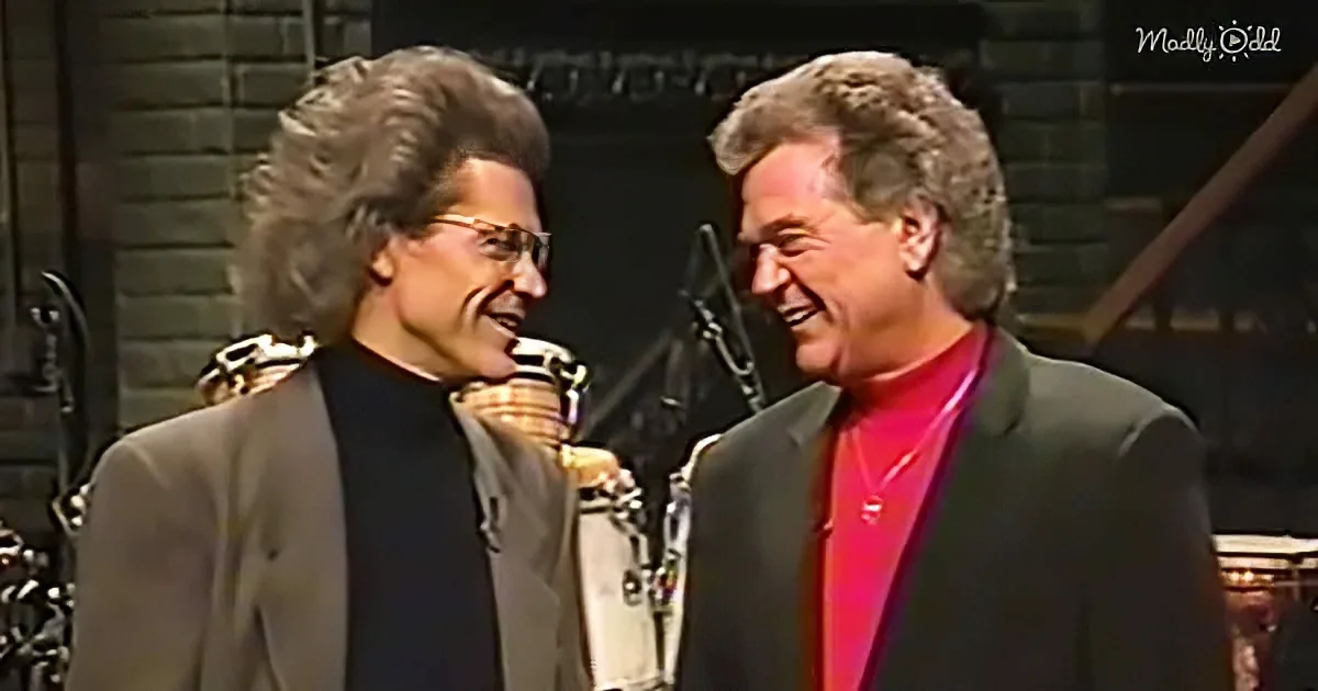 Two individuals in formal attire share a moment, smiling, with a brick wall and musical equipment in the background.
