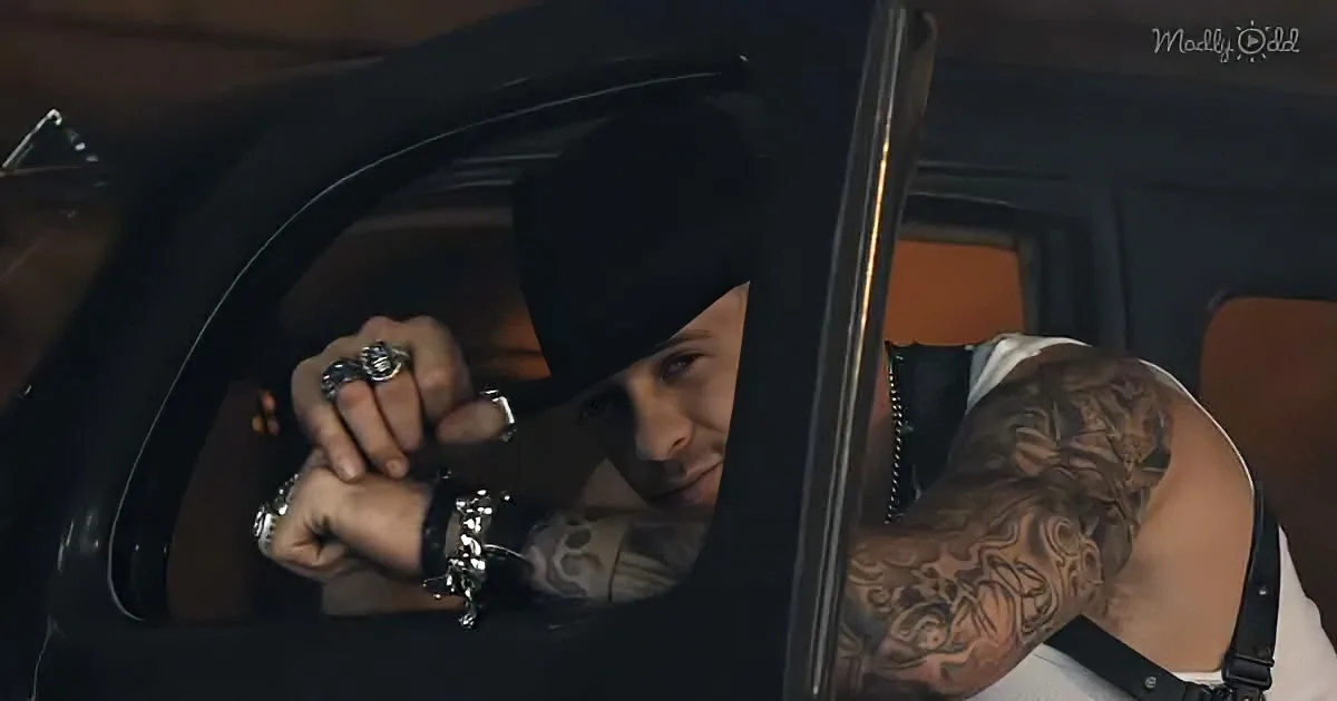 A tattooed man in a car, wearing a beanie and multiple rings, looks out the window.